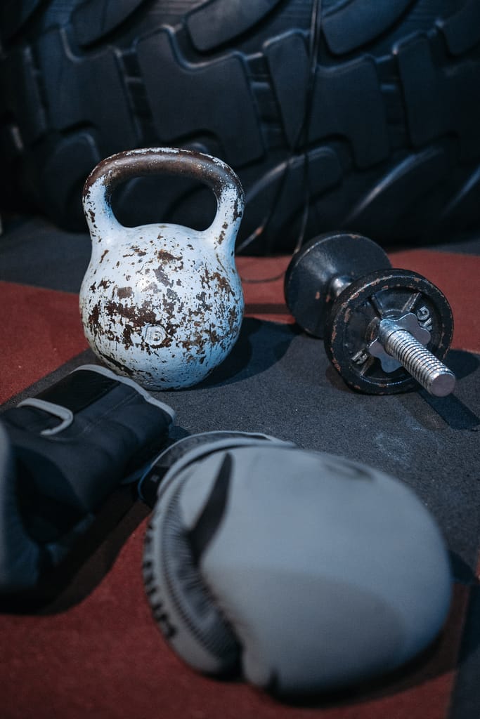 Close-up of fitness equipment including kettlebell, dumbbell, and boxing glove on gym floor.